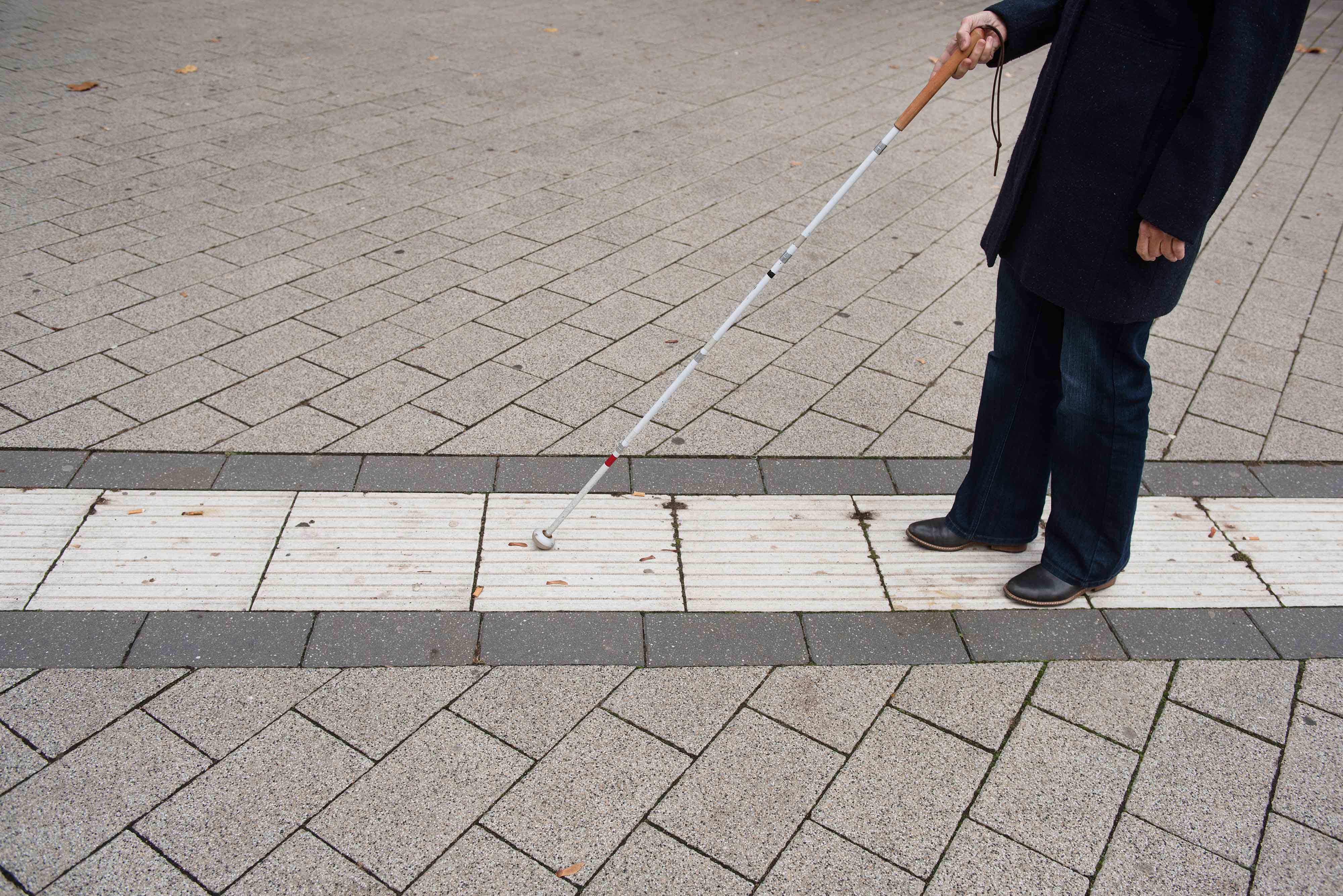 Blindenleitsystem Eine blinde Person folgt dem Blindenleitsystem auf dem Campus mithilfe eines Blindenstocks